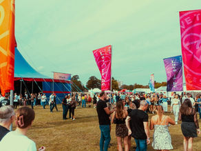 Crowd at an outdoor festival near a blue stage tent, with colorful flags reading love to be and festival under a bright sky.