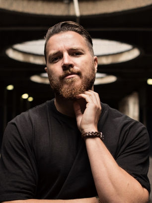 Bearded man in a black shirt poses thoughtfully in a dim tunnel with circular lights overhead.