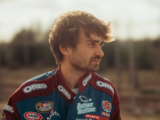 Man in a racing suit with Oreo and NASCAR logos stands outdoors, looking thoughtful. The background is blurred and sunlit.