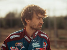 Young man in a racing suit with various logos, including Oreo, gazes right. Blurred outdoor background, giving a contemplative mood.