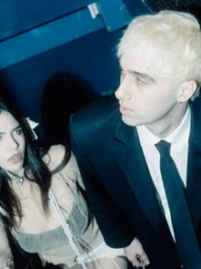 A man in a suit and a woman in a lace outfit walk up dark stairs. The setting is dimly lit with blue tones, creating a moody ambiance.