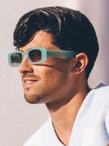 Man with short dark hair wearing teal sunglasses and a white sleeveless shirt, showing a tattooed arm, smiles in bright daylight.