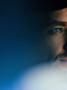 Man with blue eyes and mustache in a cap, looking thoughtful. Dim, cool-toned lighting creates a serene, introspective mood.