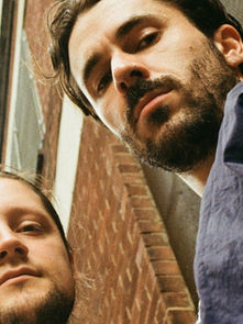 Two men looking down towards the camera with serious expressions, wearing casual jackets. Brick wall in background suggests urban setting.
