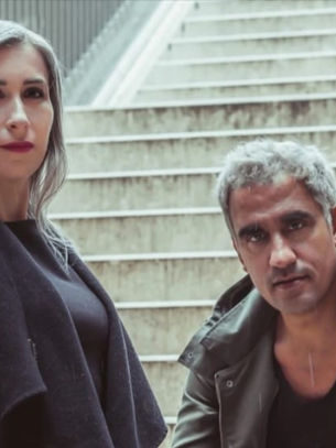 A woman stands and a man crouches on stairs, both with neutral expressions. Background of metal rails and beige steps, creating a modern urban vibe.