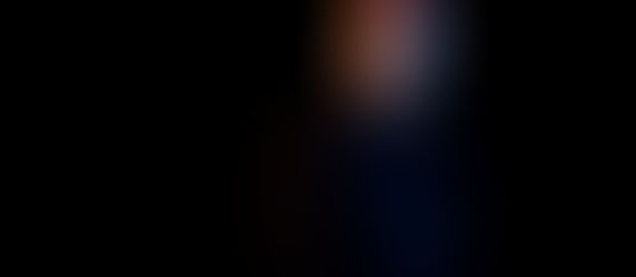 Bald man in a dark jacket, highlighted by blue and orange lighting, looks to the side against a black background. Mood is contemplative.