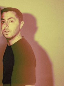 Man in black shirt stands against a yellowish wall with a ghostly shadow effect. The mood is mysterious and atmospheric.