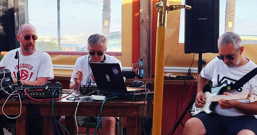 Three men in sunglasses perform music at a seaside bar, using a laptop, mixer, and guitar; Bose speaker and band T-shirts visible.