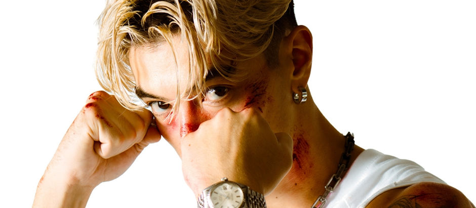 Blonde man in white tank top with clenched fists, appearing confident. He has a watch, tattoos, and slight bruises, set against a white background.