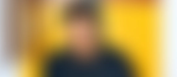 Man in glasses and blue shirt poses against a vibrant yellow and brown abstract background, conveying a thoughtful mood.