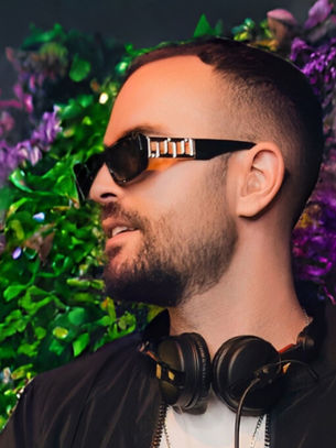 Man with sunglasses and headphones in front of vibrant green and purple floral background. Happy expression, wearing a black jacket.