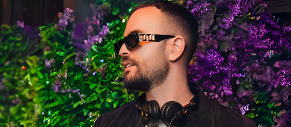 Man with sunglasses and headphones in front of vibrant green and purple floral background. Happy expression, wearing a black jacket.