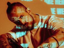 Man with tattoos leans forward, blue and white text projections on orange background. Mood is contemplative. Text partially visible.