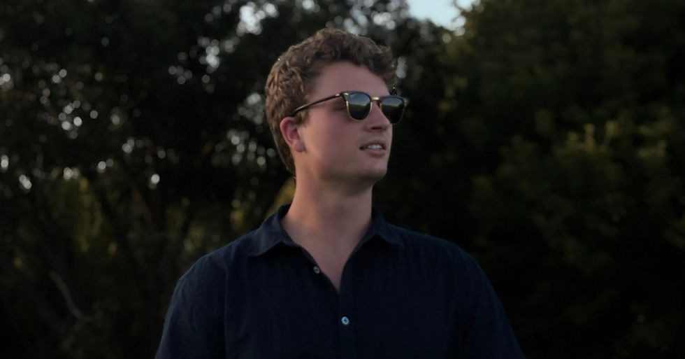 Young man in sunglasses and a dark navy shirt looks off to the right outdoors against leafy trees.
