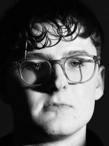 Moody black-and-white portrait of a person with glasses and curly hair, looking serious. Dark background emphasizes their expression.