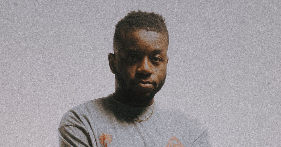 Man in a gray shirt with orange design stands against a plain gray background, looking serious.