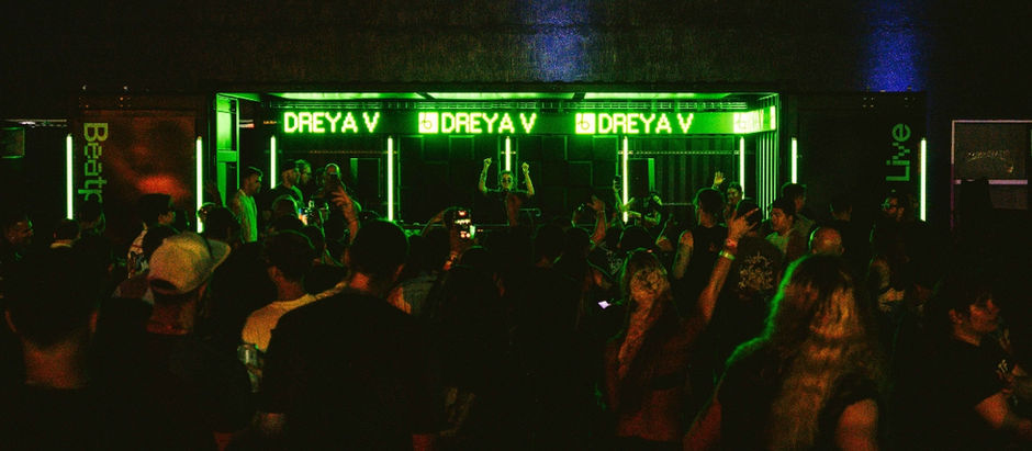 Crowd enjoying a DJ performance under green neon lights with the name "DREYA V" displayed. People are dancing and taking photos.