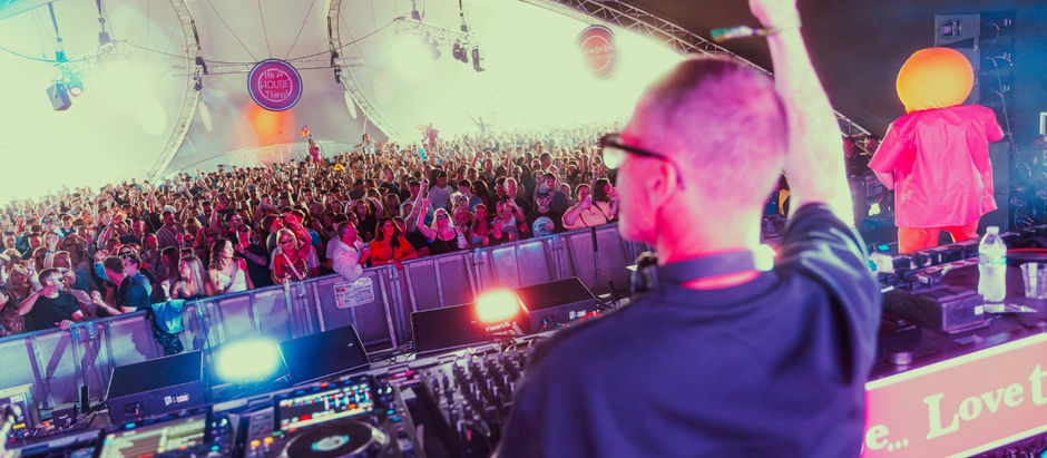 DJ playing to a lively crowd in a large tent. Colorful lights, vibrant atmosphere. "It's A House Thing" sign visible above crowd.