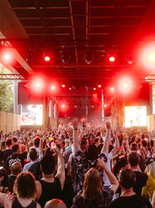 Crowd at an outdoor concert under a canopy with bright red lights. People are raising hands, showcasing excitement and energy.