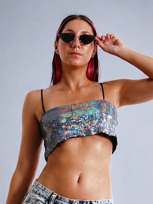 Woman in sunglasses adjusts them, wearing a shimmering crop top and jeans against a plain gray backdrop, confident pose.