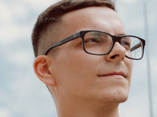 Man with black glasses looks pensively into the distance against a cloudy sky, reflecting a calm, thoughtful mood.