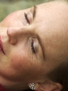 Woman in a red top resting with closed eyes, lying outdoors on grass. She wears pearl earrings and appears relaxed.
