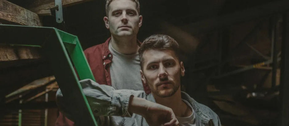 Two men stand in a dim attic, one in a red shirt, the other in denim, leaning on a green ladder. Mood is serious, no visible text.