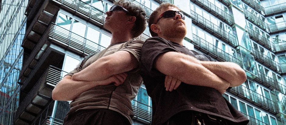 Two men in sunglasses stand back to back with arms crossed in front of a modern glass building.