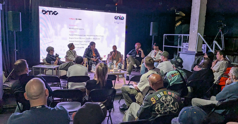 Panel discussion with seven people on stage, audience seated around. Large screen displays event details. Dimly lit, relaxed atmosphere.