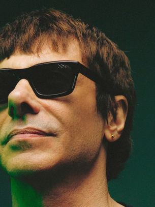 Close-up of a person in black sunglasses and green light against a dark background, looking serious.