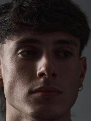 Young man with short dark hair and an earring gazes to the side in a dimly lit setting. The mood is contemplative and subdued.