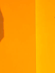 Man in black jacket walks against a vibrant orange background. Shadows of figures and hands appear on the wall, adding intrigue.