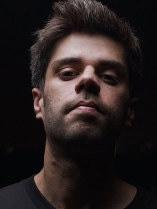 Man with serious expression in a dark setting, illuminated against a backdrop of blurred city lights. Wearing a black shirt.