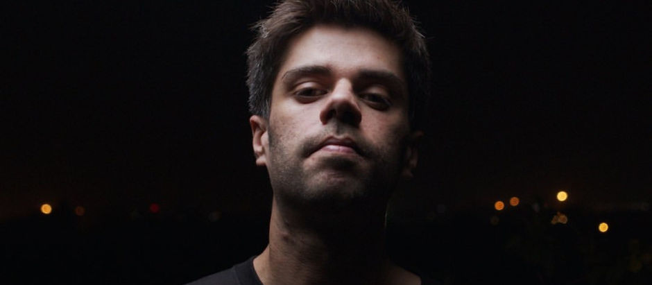Man with serious expression in a dark setting, illuminated against a backdrop of blurred city lights. Wearing a black shirt.