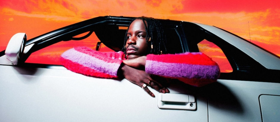 Person in a pink and purple sweater leans on a white car door, gazing thoughtfully. Vivid orange sky in the background.