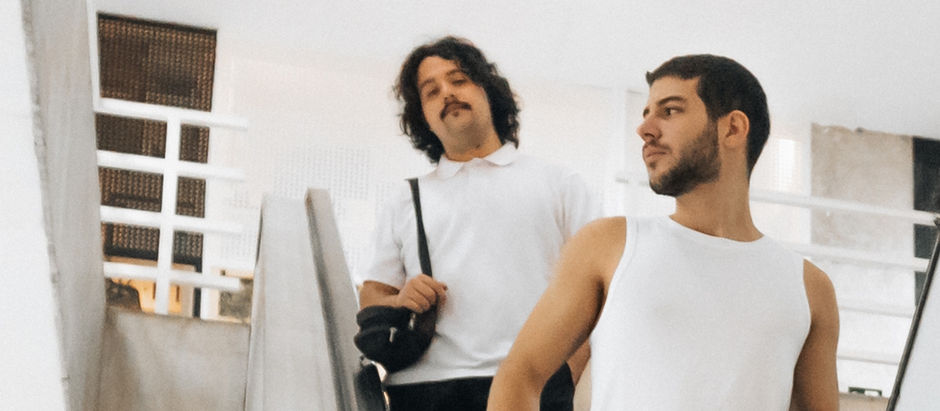 Two men on escalator; one in a white polo holds a bag, the other in a white tank top. Bright, minimalist interior. Relaxed mood.