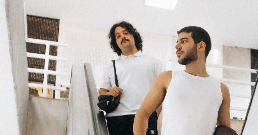 Two men on escalator; one in a white polo holds a bag, the other in a white tank top. Bright, minimalist interior. Relaxed mood.
