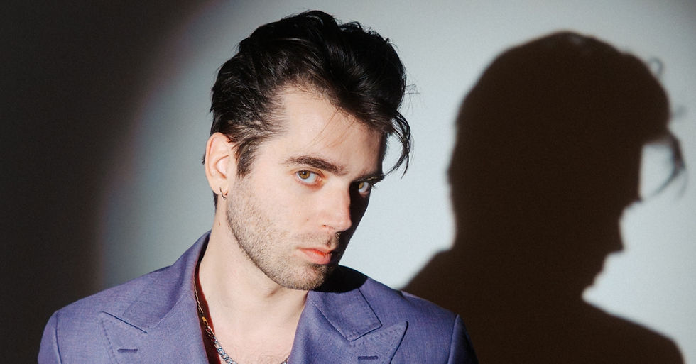 Man in purple jacket gazes intensely at camera. Strong shadow cast on wall in minimalistic setting. Mood is dramatic and mysterious.