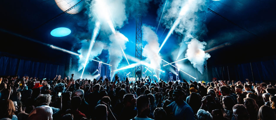 Crowded concert under blue stage lights and smoke, with people raising hands in a high-energy indoor tent.
