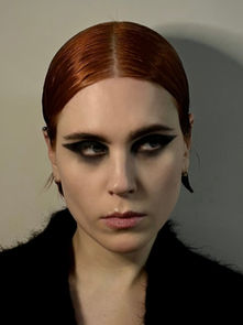Portrait of a red-haired woman with bold black eye makeup and a black coat, staring sideways against a plain gray wall.