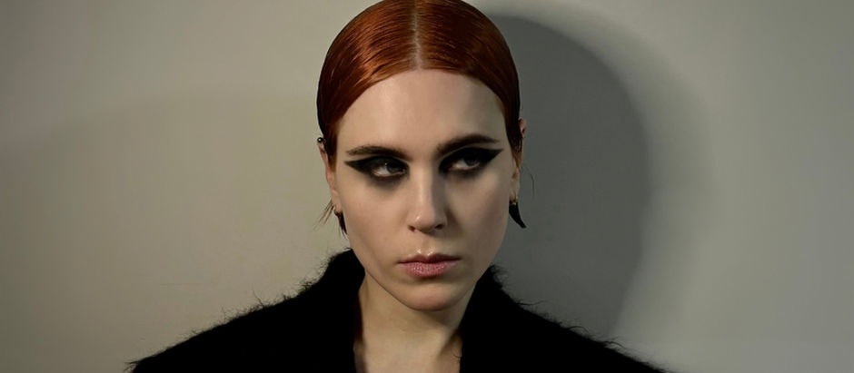 Portrait of a red-haired woman with bold black eye makeup and a black coat, staring sideways against a plain gray wall.