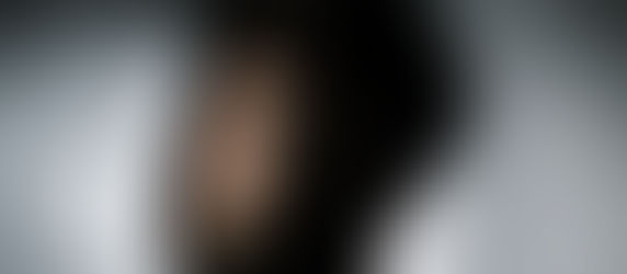 Man with beard in shadowy light, gazing at the camera. Soft focus, gray background, moody atmosphere.