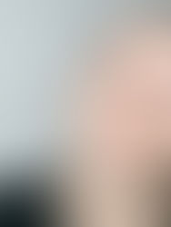 Person with short hair in a black shirt, looking into the camera with a neutral expression. Gray background and shadow.