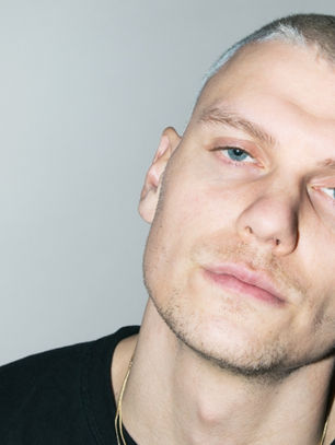 Person with short hair in a black shirt, looking into the camera with a neutral expression. Gray background and shadow.