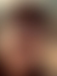 Man in a black cap and shirt gazes at the camera, standing in a dry, mountainous landscape. The background shows a forested valley.