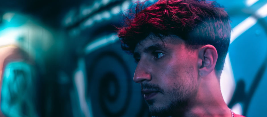 Close-up of a man in neon blue and pink light against a blurred graffiti wall, looking pensive.
