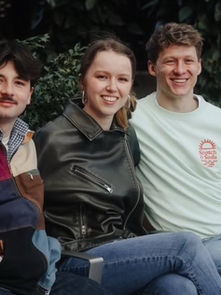 Three people sit on a bench outdoors, smiling. One wears a colorful jacket, another a leather jacket, and the third a mint sweatshirt.