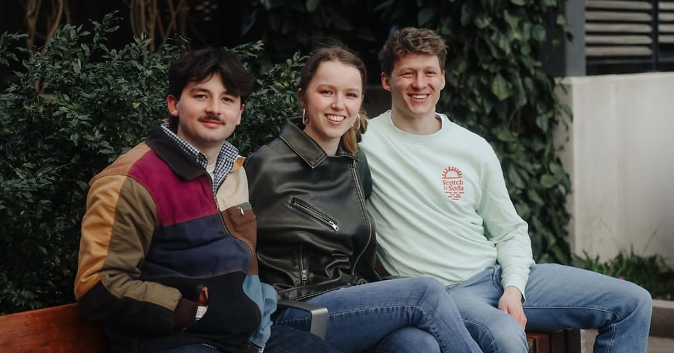 Three people sit on a bench outdoors, smiling. One wears a colorful jacket, another a leather jacket, and the third a mint sweatshirt.