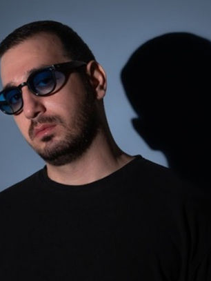 Man in sunglasses wearing a black shirt against a blue background. Shadow cast on the wall, creating a dramatic, moody effect.