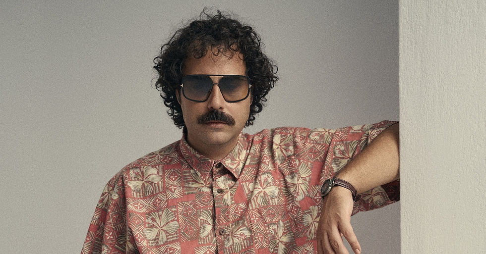 Man with curly hair, mustache, and sunglasses leans on a white wall, wearing a red patterned shirt against a gray backdrop.